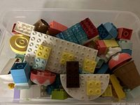 Top-down view of a container filled with 50+ multicolor LEGO Duplo pieces including baseplates, translucent blocks, and themed ice cream and cupcake shapes.