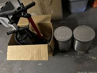 Four manual pumps in a cardboard box next to two stainless steel small step trash cans on a concrete floor.