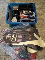 Blue plastic bin containing folded costume pieces, decorations and packaged items including a fabric skeleton hood piece and flat decorations.