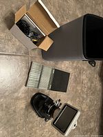 Photo showing the microscope with its LCD screen, a box with cables, multiple glass slides in a storage holder, and gray carrying case on the floor.