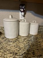 Three white ceramic canisters with lids on kitchen counter, front view showing size difference.