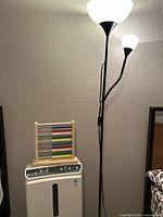 Black metal floor lamp with three varying sized frosted glass shades, electric air purifier below it, and wooden abacus on top of the purifier against a wall.