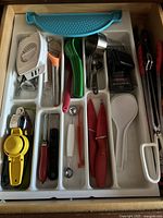 Overhead view of kitchen utensils in drawer organizer including peelers, measuring spoons, citrus juicer, egg slicer, knives, tongs and rice paddle in multicolor set.