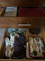View of kitchen drawer with oven mitts, potholders, and kitchen towels neatly stored. Owl and floral patterns are clearly visible on several items.