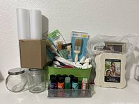 Wide view of lot showing acrylic paint bottles, paintbrushes in green container, two photo frames, glass jars, rolled canvases or paper, and crafting accessories.