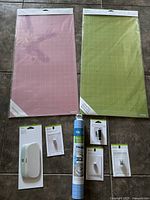 Two Cricut cutting mats in pink and green, each 12x24 inches, plus multiple Cricut branded accessories including pens, blades, and tools on floor surface.