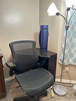 Office chair with black mesh seat and back, adjustable armrests and wheels, next to black corner desk and blue yoga mat in corner