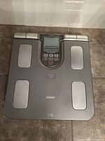 Top-down view of gray Omron HBF-514C body composition monitor showing four silver electrode pads, display screen, and control buttons, placed on tiled floor.