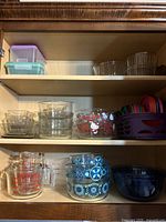 Wide view of three shelves showing clear glass Pyrex products, small plastic containers with lids on top shelf, and colorful plastic lids stored in a purple basket on middle shelf.