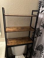 Full view of the rustic freestanding over toilet shelf, showing three stained wooden shelves framed in black metal, placed over a white toilet.