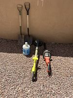 Photo showing two metal shovels with wooden handles, electric weed wacker, electric trimmer, and plastic garden sprayer container set on gravel background.