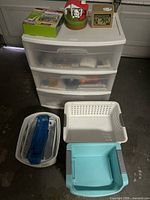 Sterilite three-drawer plastic storage unit with three semi-translucent drawers holding various items. Four plastic baskets and trays in white, blue, and teal arranged in front of drawers.