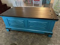 View of teal wooden storage chest showing black plank top and carved front panels.