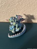 Front view of a small Tesori porcelain dragon figurine holding a blue Swarovski crystal, painted in green, blue and pink hues.