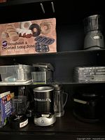 Overview of Nutribullet blender with multiple cups, Hamilton Beach grinder, Wilton baking set box, and assorted glass/plastic bakeware stack on dark shelf