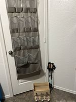Full view of fabric over-the-door organizer with clear pockets hanging on a white door, two walking sticks leaning against the wall, a small wooden box and owl wall hooks on the floor.