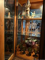 Glass cabinet with fabric elephants on the top shelf, crystal glasses in the middle, and green decorative glass goblets on the bottom shelf with artificial grapes in a purple bowl.