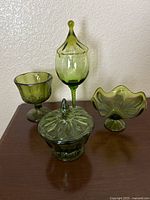 View of all four pieces of green glassware set arranged on a surface: candy dish with lid, goblet, tall goblet with twisted stem, and ruffled pedestal compote bowl.