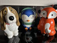 Three ceramic cookie jars shaped as a dog, penguin with earmuffs, and fox on a stove top.