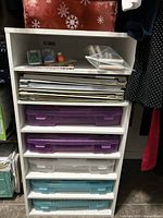 White composite wood shelving unit with five plastic drawers and stacked paper