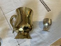Close-up of a single silvered beer mug with handle alongside a silvered brandy shot glass, showing engraved floral designs and polished finish.