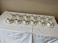 Set of 12 coffee/tea cups with matching saucers arranged on a white embroidered tablecloth, showing all pieces in good condition and unused.