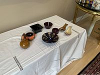 Photo of colorful pottery pieces including a small brown round vase, square black dish, lidded orange-brown bowl, blue bowl with tan spots, purple bowls, and two decorative spoon holders placed on a white tablecloth.