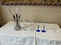 6 crystal champagne glasses with different colored stems in a clear jar, 2 clear stem glasses, and 2 blue stem GeoQuest glasses on a white tablecloth.