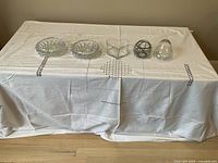 Five assorted small glass bowls arranged on a table covered with white cloth, including two round clear pressed glass bowls, one heart-shaped clear glass bowl, one round glass bowl with black wire design, and one dome-shaped clear glass bowl.