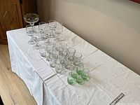 Front and side view of all glasses arranged on a white cloth-covered table showing clear and green glasses including margarita and Irish coffee glasses.