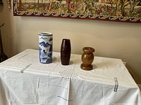 Three vases placed on a white embroidered cloth against a neutral background, showing the relative sizes and styles: a blue and white porcelain vase, a dark brown smooth vase, and a wooden rounded vase.
