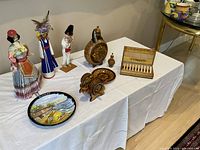 Photo showing entire assortment displayed on white tablecloth including the three dolls, bottles, wooden items, cigarette case, plate and painting