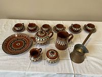 Full set of Bulgarian pottery coffee and tea items arranged on a white cloth. Includes 6 small coffee cups with saucers, a tea pot, coffee pot, sugar and milk containers, decorative plate, and a copper Turkish coffee pot.