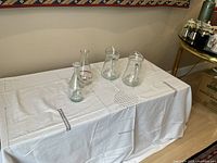 Four glass carafes displayed on a white cloth-covered table against a wall, showing all items in the lot.