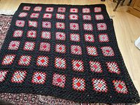 Full view of crocheted afghan showing black grid and multicolored squares in red, gray, orange, beige tones on dark background spread on floor.
