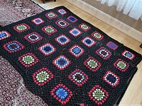 Image showing the full laid out hand crocheted afghan featuring a black background with colorful multi-colored squares in red, white, green, and blue patterns.
