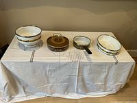 Front view of the full set including three ovenproof porcelain and stainless steel dishes and one large porcelain sieve with metal stand on white tablecloth.
