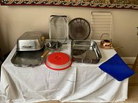 Full view of all kitchen articles laid out on table including roasting pan, trays, sieve, brass water can, rubber cover, grille, and masher.