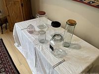 Multiple kitchen jars including glass and plastic on a white cloth-covered table, showing variety of jar sizes and lids.
