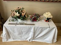 Three silk flower arrangements on a draped table topped with white cloth in front of a wall, with a measuring stick showing sizes.