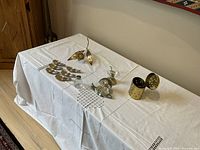 Wide view of six silvered decorative items on a white embroidered cloth, includes flower, snail butter dish, salt dispenser, napkin holder, jam jar, and carved box.