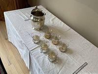 Photo showing silvered metal carafe and six glass cups with silvered holders arranged in row on a white embroidered tablecloth.