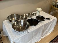 Four stainless steel bowls of varying sizes, large sieve with legs, small kitchen scale, and two non-stick pans displayed on a table.