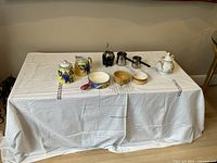 Photo showing all main tea and coffee items on a table covered with a white cloth: Japanese teapot, sugar and milk set with grape motif, three dishes of various types, Argentine tea cup, and two metal Turkish coffee pots.