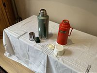 Photo showing two thermoses (green stainless steel and orange/red glass) with their lids and stoppers laid out on a white cloth-covered table.