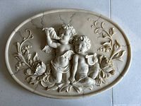 Oval angel wall hanging featuring two cherubic angels in relief with floral and leaf designs, placed on a tiled surface.