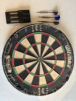 Photo showing a round regulation Winmau bristle dart board with scoring segments and numbers, three steel tip darts with flights, and a black plastic dart holder.