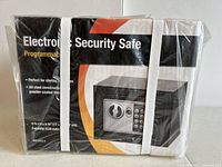 Photo shows the unopened Electronic Security Safe in its original packaging with plastic wrap. The box states it is a programmable keypad safe with steel construction and powder-coated finish, size about 6.75 x 5 x 6.75 inches.