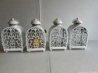 Four white metal candle lanterns with glass panels and decorative scrollwork, seen from the front on a neutral background.