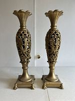Front view of two gold-tone metal candlesticks with intricate floral and leaf cutout designs and square claw foot bases.